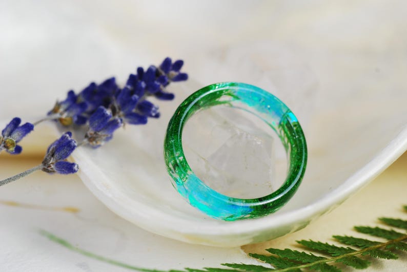 Op de afbeelding: Een groene en blauwe harsring met ingebedde botanische elementen. De ring ligt op een wit oppervlak met lavendelbloemen en groene bladeren.