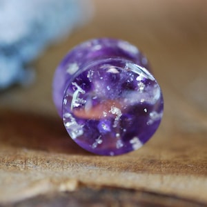 Puede incluir: Primer plano de un expansor de oreja morado y blanco con un acabado brillante. El expansor est&aacute; hecho de resina y tiene una apariencia clara y transl&uacute;cida.