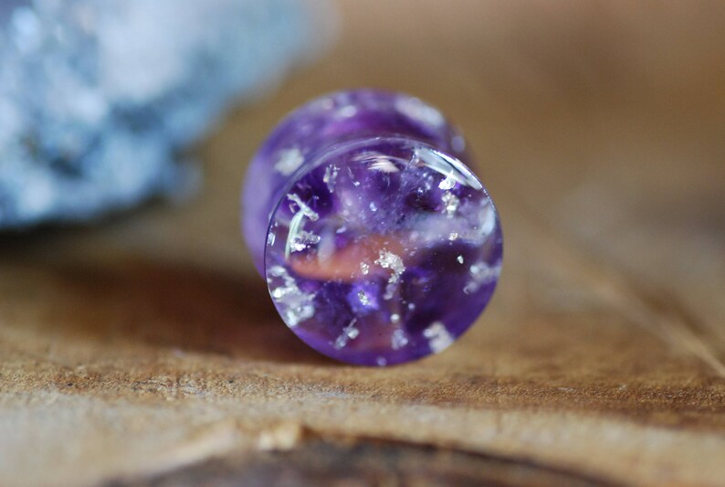 Puede incluir: Primer plano de un expansor de oreja morado y blanco con un acabado brillante. El expansor est&aacute; hecho de resina y tiene una apariencia clara y transl&uacute;cida.