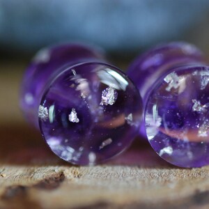 Puede incluir: Dos tapones de vidrio morado con motas plateadas. Los tapones son redondos y tienen una superficie lisa y brillante.