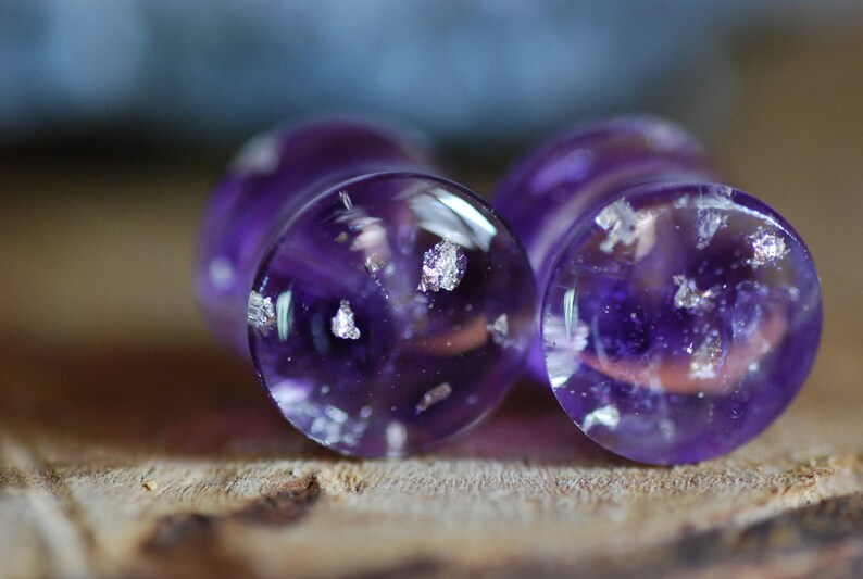 Puede incluir: Dos tapones de vidrio morado con motas plateadas. Los tapones son redondos y tienen una superficie lisa y brillante.