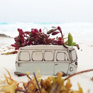 Peut inclure: Un pot en céramique blanche en forme de van vintage avec des succulentes rouges et vertes qui poussent sur le dessus. Le pot est posé sur une plage de sable blanc.