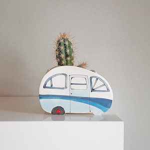 Peut inclure: Pot en céramique blanche en forme de camping-car vintage avec des détails bleus et gris. Une petite plante de cactus pousse à l'intérieur du pot.