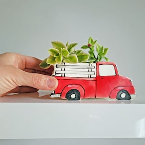 Peut inclure: Planteur en forme de camion rouge en céramique avec des accents blancs et une benne à lattes de bois. Le camion est rempli de succulentes vertes.