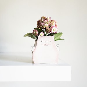 Peut inclure: Un pot en céramique rose en forme de cochon avec un visage souriant et des ailes. Le pot est rempli de fleurs roses et blanches.