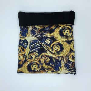 May include: A neck warmer with a black fleece top and a patterned bottom. The pattern features a swirling design in shades of yellow, blue, and brown, with a depiction of the TARDIS from Doctor Who. The neck warmer is designed to be folded over.
