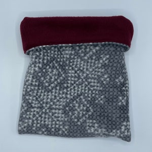 May include: A fleece bag with a gray and white patterned body and a folded burgundy top. The bag is made of soft material and is likely used for storage or as a gift bag. The pattern is a repeating geometric design.