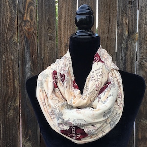 May include: Cream-colored infinity scarf with a map design in burgundy and black. The scarf is draped on a black mannequin. The map design includes text and illustrations. The background is a wooden fence.