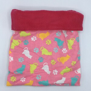 Puede incluir: Una cama rosa para mascotas con una parte superior de forro polar roja. La cama presenta un patrón de siluetas de gatos coloridos y huellas de patas. La cama está diseñada para mascotas pequeñas.