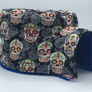 May include: A decorative, padded item featuring a black fabric with a repeating pattern of colorful sugar skulls and gray floral accents. The item has a blue trim and a blue interior.