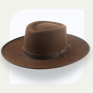 May include: A brown fedora-style hat with a wide brim. The hat features a dark brown leather band secured with a small gold-colored buckle. The hat's crown is rounded, and the brim is slightly upturned.
