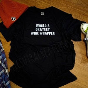 May include: Black t-shirt with the text "WORLD'S OKAYEST WIRE WRAPPER" in white. A red beanie with a white and black logo, a blue plaid shirt, and a pair of neon green sneakers with checkered laces are also in the image.