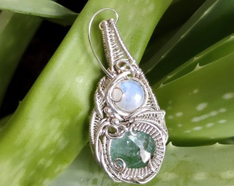Può includere: Un ciondolo avvolto in filo d'argento con una pietra preziosa verde e bianca. Il ciondolo è mostrato su uno sfondo di pianta verde.
