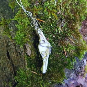May include: A white bone pendant wrapped in silver wire, hanging from a silver chain. The pendant is shaped like a bird's wing and has a spiral design at the top. The pendant is resting on a bed of moss.