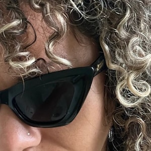 May include: A person with curly blonde hair wearing black cat-eye sunglasses with a small nose ring.