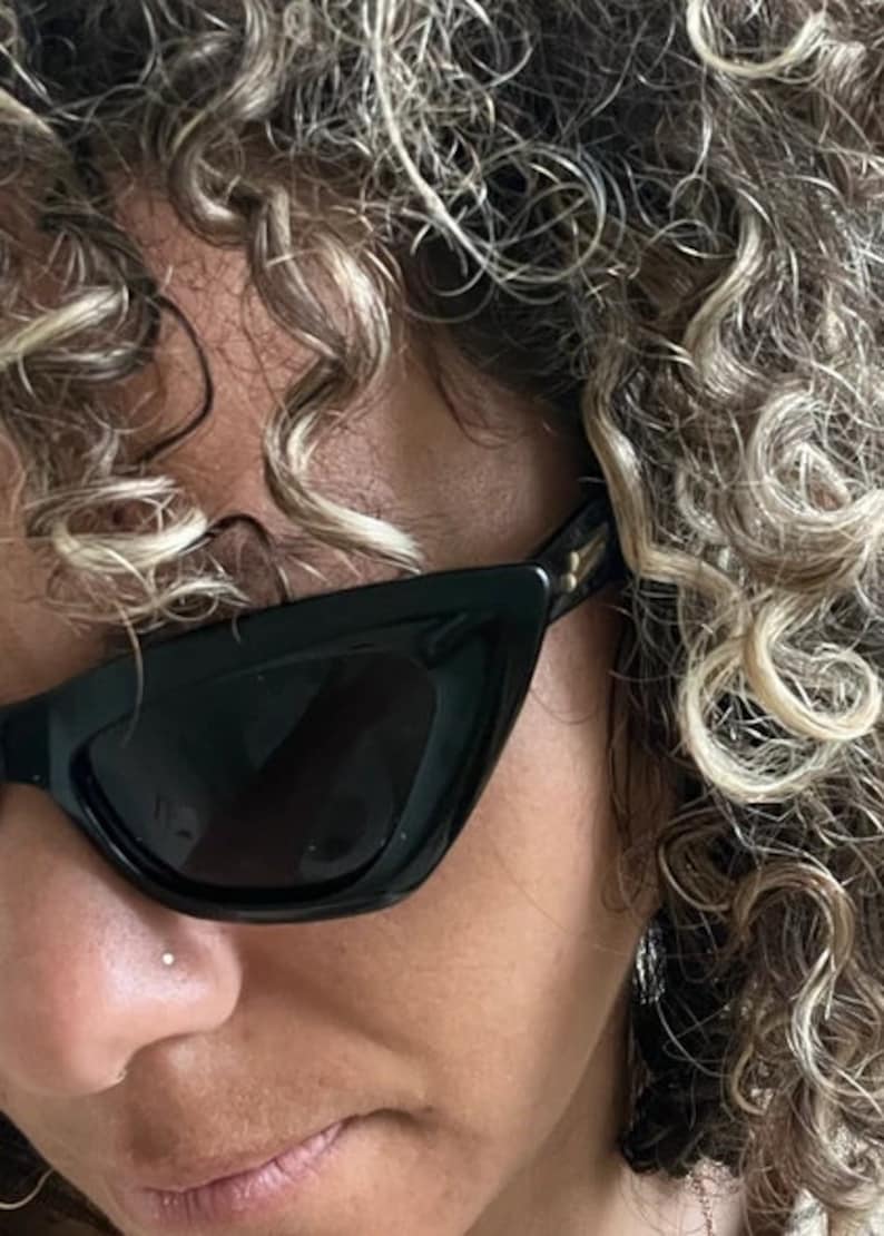 May include: A person with curly blonde hair wearing black cat-eye sunglasses with a small nose ring.