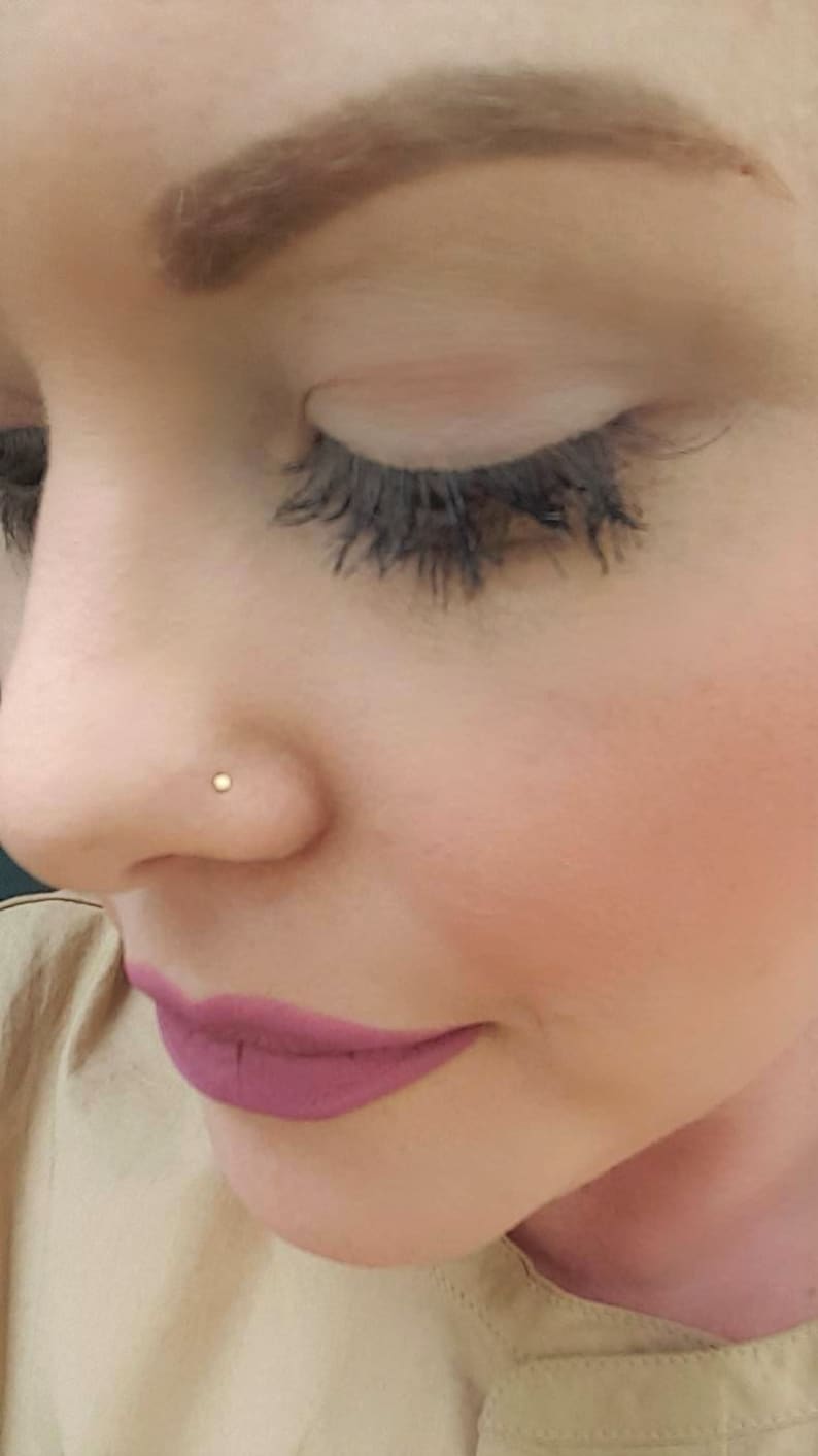 May include: Close-up of a person's face with a small gold nose ring. The person has long eyelashes and is wearing pink lipstick.