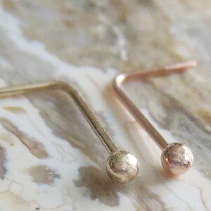 May include: Two gold nose rings with a small ball at the end. The rings are bent at a 90-degree angle.