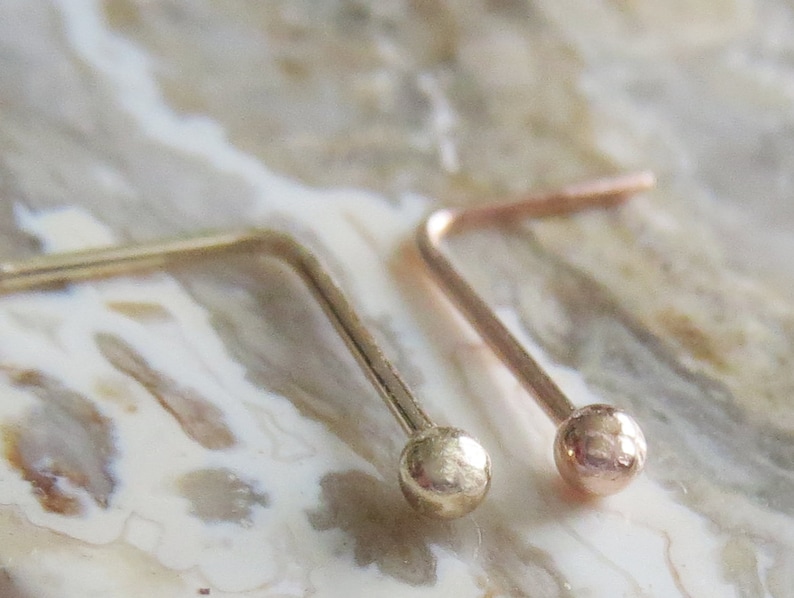May include: Two gold nose rings with a small ball at the end. The rings are bent at a 90-degree angle.