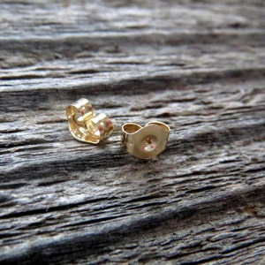 May include: Two gold-colored earring backs, also known as butterfly backs, are shown on a wooden surface. The backs are made of metal and have a rounded shape.
