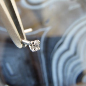 May include: A small, clear, round gemstone set in a silver setting. The gemstone is held by tweezers against a background of white and gray agate.