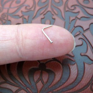May include: A silver nose ring with a small L-shaped bend. The ring is held between a finger and thumb.