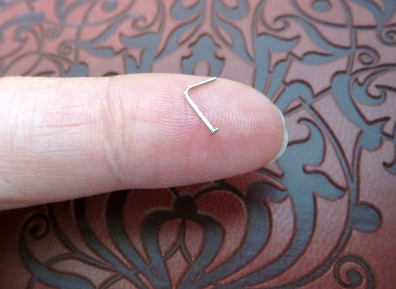 May include: A silver nose ring with a small L-shaped bend. The ring is held between a finger and thumb.