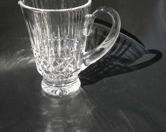 May include: A clear crystal pitcher with a handle and a pouring spout. The pitcher has vertical and diamond-shaped cut glass patterns. The pitcher sits on a round base. The background is dark, and the light creates reflections.