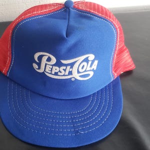 Op de afbeelding: Een blauwe en rode Pepsi-Cola truckerpet. De pet heeft een blauw voorpaneel met het Pepsi-Cola-logo in witte letters. De mesh achterkant is rood en de klep blauw met witte stiksels.