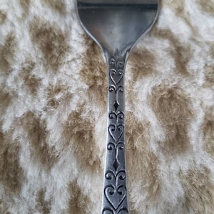 Puede incluir: Primer plano de un tenedor plateado con un mango decorativo. El mango presenta un patrón grabado de corazones y diamantes. Las púas son visibles en la parte superior. El tenedor está sobre un fondo texturizado de color marrón claro.