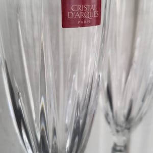 May include: Two clear crystal wine glasses with a textured, cut-glass design. A red label on one glass reads "Cristal d'Arques PARIS". The glasses have a long stem and a wide bowl, ideal for serving wine.