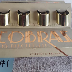 Peut inclure: Un ensemble de quatre ronds de serviette en métal doré dans une boîte. La boîte est beige avec le mot "COBRAS" en lettres dorées. Les mots "COPPER & BRASS" sont également visibles sur la boîte.
