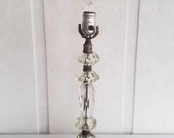 May include: A crystal table lamp with a clear glass base and a clear glass shade. The lamp has a metal base and a metal shade holder. The lamp is on a white surface.