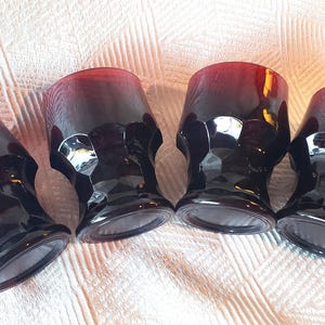 May include: Four dark red and black faceted drinking glasses. The glasses have a gradient color, transitioning from red at the top to black at the base. The glasses are arranged on a white textured surface.