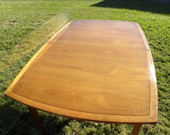 May include: A rectangular wooden table with a light brown finish and rounded corners. The table has a glass top and is set on a grassy surface. The table measures approximately 60 inches long and 36 inches wide.