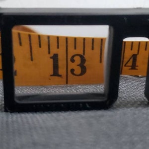 May include: Close-up of a yellow measuring tape with black markings, showing the numbers 11, 12, 13, 14, and 15 inches. A black frame is in the foreground, partially obscuring the tape. The background is a gray fabric.