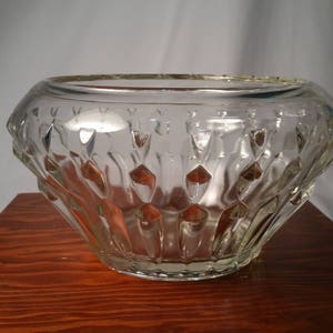 May include: A clear glass bowl with a diamond-patterned exterior. The bowl has a wide, rounded shape and sits on a wooden surface. The glass is transparent, allowing light to pass through.