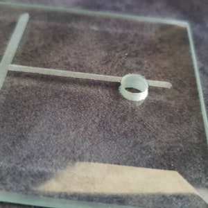 May include: A clear glass object with etched lines forming a cross and a small circular detail. The glass is rectangular with a beveled edge. The background is a dark, textured surface.