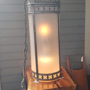 May include: A hanging lantern with a frosted glass cylinder, framed by ornate metalwork. The lantern is suspended by a chain and sits on a wooden table. The metalwork features decorative patterns and a crown-like top.