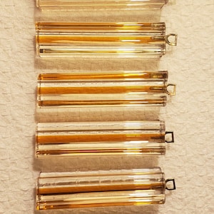 May include: Five clear glass tubes with a golden-yellow interior and a small, rectangular metal handle at one end. The tubes are arranged vertically against a textured, off-white background.