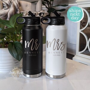 May include: Two stainless steel water bottles with black and white finishes. The bottles have a black lid with a loop handle and a straw. The bottles are personalized with the text "Mr. Bennett 10.22.2022" and "Mrs. Bennett 10.22.2022".
