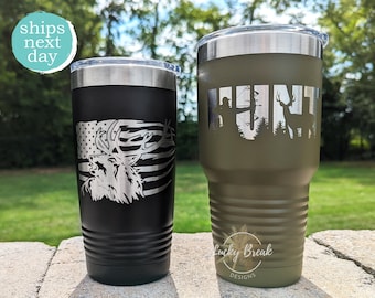 Puede incluir: Dos vasos de acero inoxidable con diseños de temática de caza. El vaso negro presenta una cabeza de ciervo con un fondo de bandera estadounidense. El vaso verde presenta la palabra "HUNT" con una silueta de un cazador y un ciervo en el bosque.