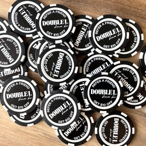 Op de afbeelding: Een verzameling zwart-witte promotiechips met de tekst "DOUBLEL" en "REFER A FRIEND GET $10 OFF". De chips liggen verspreid op een lichtbruin houten oppervlak.