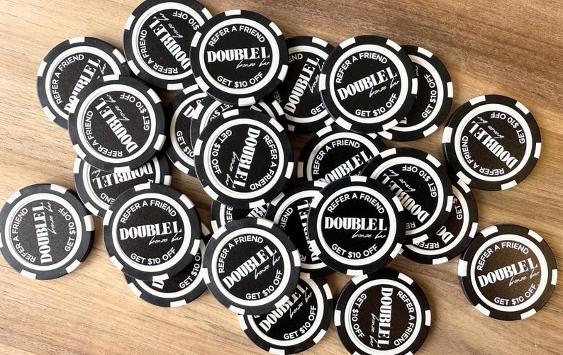 Op de afbeelding: Een verzameling zwart-witte promotiechips met de tekst "DOUBLEL" en "REFER A FRIEND GET $10 OFF". De chips liggen verspreid op een lichtbruin houten oppervlak.