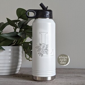 May include: White stainless steel water bottle with a black lid and a floral design featuring the letter L.