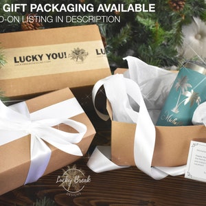 May include: Gift set with a teal tumbler featuring palm tree designs and a personalized name, nestled in a kraft paper box with white ribbon. The box is inside a larger gift box with a white ribbon. The image includes a holiday card.