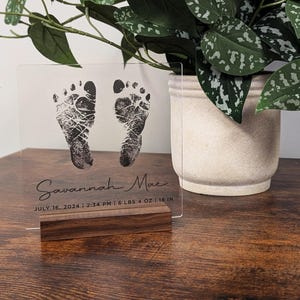 May include: A clear acrylic plaque with a wooden base, featuring two black baby footprints and the text "Savannah Mae JULY 16, 2024 | 2:34 PM | 6 LBS 4 OZ | 18 IN".
