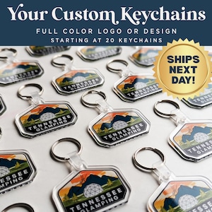 May include: A close-up of several custom keychains with a colorful design featuring a geodesic dome and the text "Tennessee Glamping". The keychains are made of clear acrylic and have a silver metal ring.