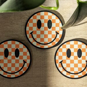 May include: Three orange and white checkered smiley face coasters. The coasters are round and have a black outline.
