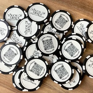 Op de afbeelding: Een verzameling zwart-witte pokerchips met de tekst "Touch of Grace" en "Verwijs een vriend voor $25 korting". De chips hebben een cirkelvormig ontwerp met een zwart-wit gestreepte rand.